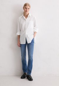 Witte blouse met opgerolde mouwen, enkele borstzak; gecombineerd met blauwe jeans en zwarte instappers. Effen achtergrond.