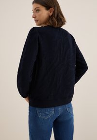 Felpa blu navy con motivo a onde testurizzato, polsini e orlo a costine. Abbinata a jeans in denim blu con tasche posteriori e cuciture.