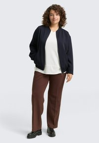 Femme aux cheveux bouclés portant une veste zippée bleu marine, un haut blanc, un pantalon marron et des bottes noires, se tenant avec une main dans la poche de la veste.