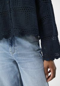Cárdigan de color azul marino, tejido a ganchillo, con cierre de botones y mangas acampanadas con bordes en forma de concha, combinado con vaqueros de mezclilla azul claro.