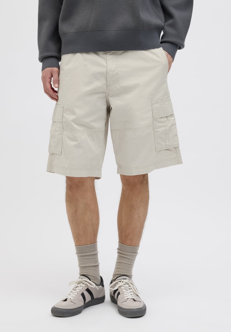 Beige Cargo-Shorts mit mehreren Seitentaschen, aus leichtem Stoff. Kombiniert mit grauen Socken und sneakers mit schwarzen Akzenten.