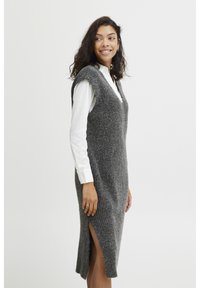 Abito grigio lavorato a maglia con scollo a V, senza maniche, fessure laterali e una finitura testurizzata, abbinato a una camicia bianca con bottoni sotto.