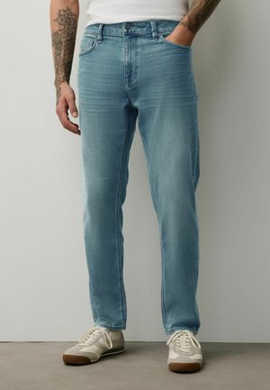 Man wearing light blue jeans and white sneakers, standing with one hand in pocket against plain background.