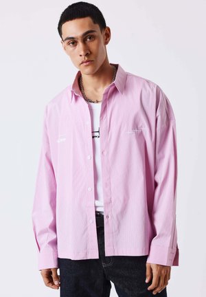 Giovane uomo con capelli neri corti che indossa una camicia rosa chiaro aperta sopra una t-shirt bianca con scritte nere e pantaloni scuri.