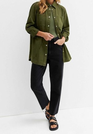 Persona con una camisa de botones verde oliva de gran tamaño, jeans negros de cintura alta con bajos sin rematar y sandalias negras con hebilla, de pie contra un fondo blanco.