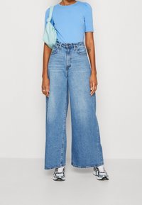 Světlomodré přiléhavé tričko s krátkými nabíranými rukávy, v kombinaci s džínami s vysokým pasem a širokými nohavicemi v modré barvě a stříbrnými teniskami. Doplňky zahrnují tyrkysovou tašku.