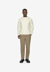 Maglione crema dalla texture morbida con scollatura rotonda, abbinato a pantaloni beige su misura e stivali neri chunky. Il modello si trova su uno sfondo neutro.