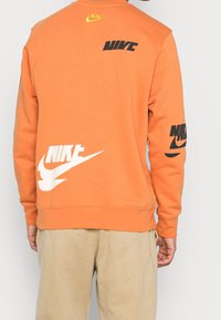 Person som bär en orange sweatshirt med flera Nike-loggor i svart, vitt och gult, kombinerat med beige byxor, sedd bakifrån.