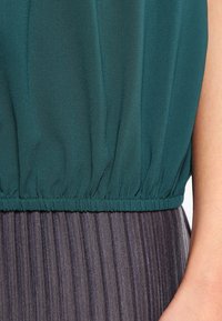 Blouse bleu sarcelle avec ourlet élastique froncé, portée sur une jupe plissée grise superposée. Caractérisée par une texture lisse et un éclat subtil.