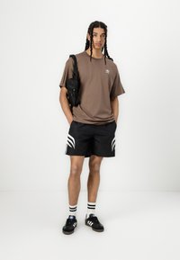 Brun bomulds t-shirt, sorte shorts med hvide striber, sorte sneakers og en lille sort crossbody-taske. Personen står med hænderne i lommen.