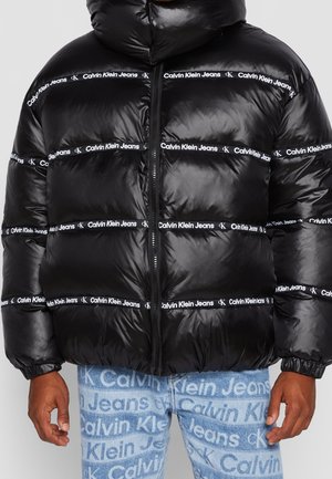 Zwarte pufferjas met horizontale witte Calvin Klein Jeans-logo strepen, gecombineerd met lichtblauwe jeans met een Calvin Klein Jeans-logo patroon.