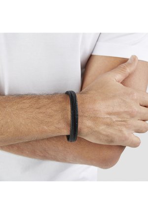Mann trägt ein schwarzes geflochtenes Lederarmband am linken Handgelenk und hält seinen rechten Ellbogen, während er ein weißes Hemd trägt.