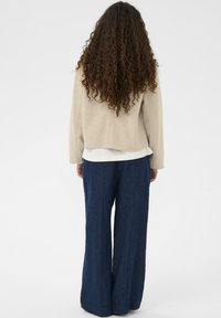 Un pull beige, coupé court, avec une texture douce, superposé sur un haut blanc ; associé à un pantalon large bleu marine avec des rayures verticales.