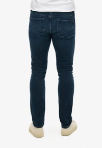 Jeans en denim bleu foncé à coupe slim, avec deux poches arrière, des coutures minimalistes et un branding discret près de la taille.