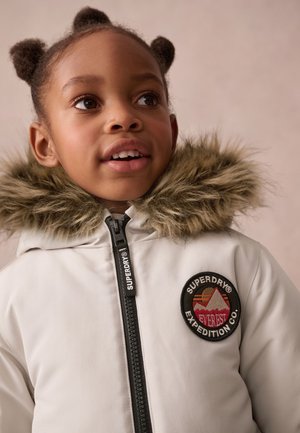 Veste blanche rembourrée avec capuche en fausse fourrure, présentant un patch logo circulaire et une fermeture éclair noire avec le marquage "SUPERDRY".