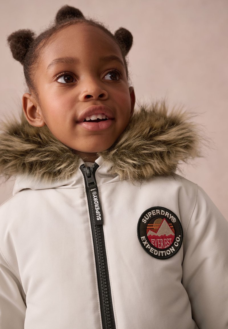 White padded jacket with a faux fur hood, featuring a circular logo patch and a black zipper with "SUPERDRY" branding.