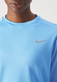 Närbild av en person som bär en ljusblå Nike-tröja med grå swoosh-logotyp på övre bröstet mot en enfärgad vit bakgrund.