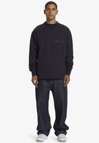 Sudadera azul marino con cuello redondo y logo texturizado "D94" en el pecho. Combinada con jeans de mezclilla oscuros de corte relax y zapatillas blancas.