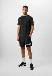 Schwarzes T-Shirt mit kurzen Ärmeln und einem verwitterten Textur, kombiniert mit schwarzen Shorts, die ein weißes "NIKE"-Grafik-Design zeigen. Das Modell trägt weiße Socken und Sneaker.