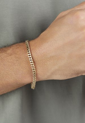Männliches Handgelenk mit dünnem Goldkettenarmband vor einem gedämpften grauen Stoffhintergrund.