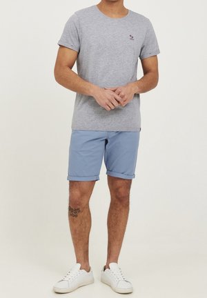 Homme portant un t-shirt à manches courtes gris clair, un short bleu retroussé, des baskets blanches, se tenant avec les mains jointes devant, tatouage sur le mollet gauche.