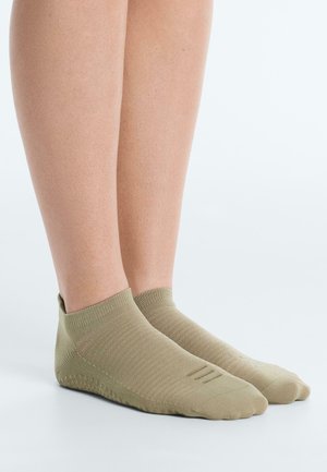 Ženské nohy v béžových ponožkách s protišmykovými bodkami na členku stoja na hladkom bielom povrchu a ukazujú textúru ponožiek a protišmykové bodky pod nimi.