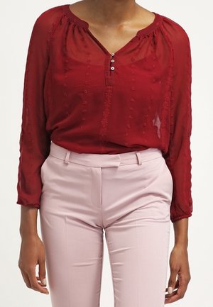Femme portant un chemisier rouge transparent à pois texturés avec trois boutons, associé à un pantalon taille haute rose clair, cintré.