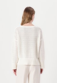 Maglione bianco lavorato a maglia con una vestibilità ampia, motivo traforato testurizzato e bordi e polsini a coste, mostrato da dietro.