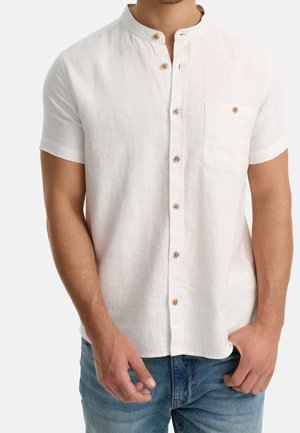 Mężczyzna ubrany w białą koszulę z krótkim rękawem, kołnierzykiem typu mandarynka i kieszonką na piersi, w połączeniu z niebieskimi dżinsami, ręce swobodnie opuszczone wzdłuż ciała.