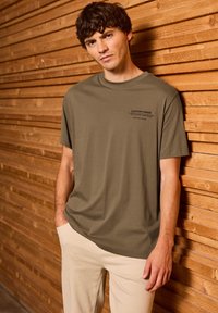 Olivově zelené tričko s krátkým rukávem a vlastním potiskem na hrudi; měkký bavlněný materiál. Model má na sobě světlé béžové kalhoty a stojí před dřevěným pozadím.