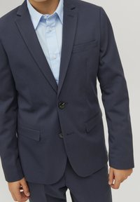 Traje azul marino con diseño estructurado, solapas con muesca, dos botones negros y una camisa de vestir azul clara debajo.