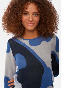 Pull coloré avec un motif de formes bleues, noires et grises. Fait en matière tricot douce avec un col rond et des manches longues.