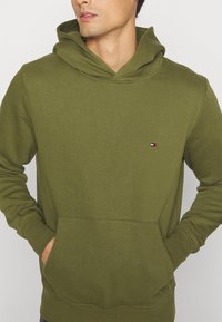 Sudadera verde olivo de tejido suave, con un bolsillo delantero, mangas largas y un parche de logo en el pecho. Diseño con capucha.