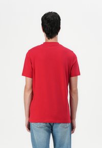 Raudona medvilnėnė marškinėliai su trumpomis rankovėmis, apvaliu iškirpimu ir reguliariai prigludimu. Nuo nugaros matoma lygi, minimalistinė dizaino koncepcija.