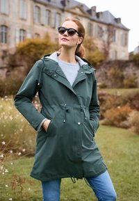 Vrouw draagt groene regenjas met kap en zonnebril, staat met handen in zakken in een tuin met een oud stenen gebouw op de achtergrond.