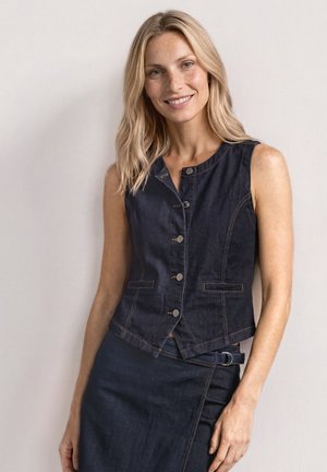 Donna sorridente con capelli biondi indossa un gilet in denim scuro senza maniche con bottoni e una gonna abbinata con dettaglio laterale con fibbia.