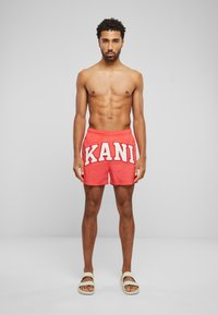 Red swimming shorts featuring white "KANI" lettering, with an elastic waistband and a relaxed fit, paired with beige sandals.