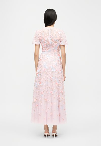 Γυναίκα με μακριά σκουρόχρωμα μαλλιά, φοράει διάφανο ροζ φόρεμα με κεντήματα από λουλούδια και λευκά σανδάλια με τακούνι, στέκεται με την πλάτη προς τα εμάς σε λευκό φόντο.