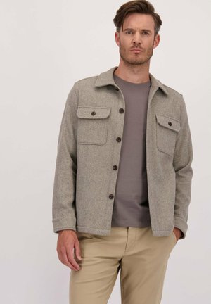 Man in een grijs herringbone knopenjasje over een grijs shirt en beige broek, staand met één hand in de zak tegen een effen achtergrond.