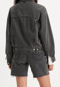 Chaqueta de mezclilla negra con un corte relajado, que presenta un cuello clásico, frente abotonado y yugo en la espalda. Combinada con pantalones cortos de mezclilla negra deshilachados.