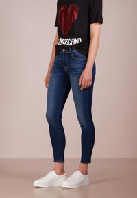 Jeans ajustados en mezclilla azul oscuro con diseño de cinco bolsillos, combinados con una camiseta negra con un corazón de lentejuelas rojas y letras blancas. Zapatillas blancas.