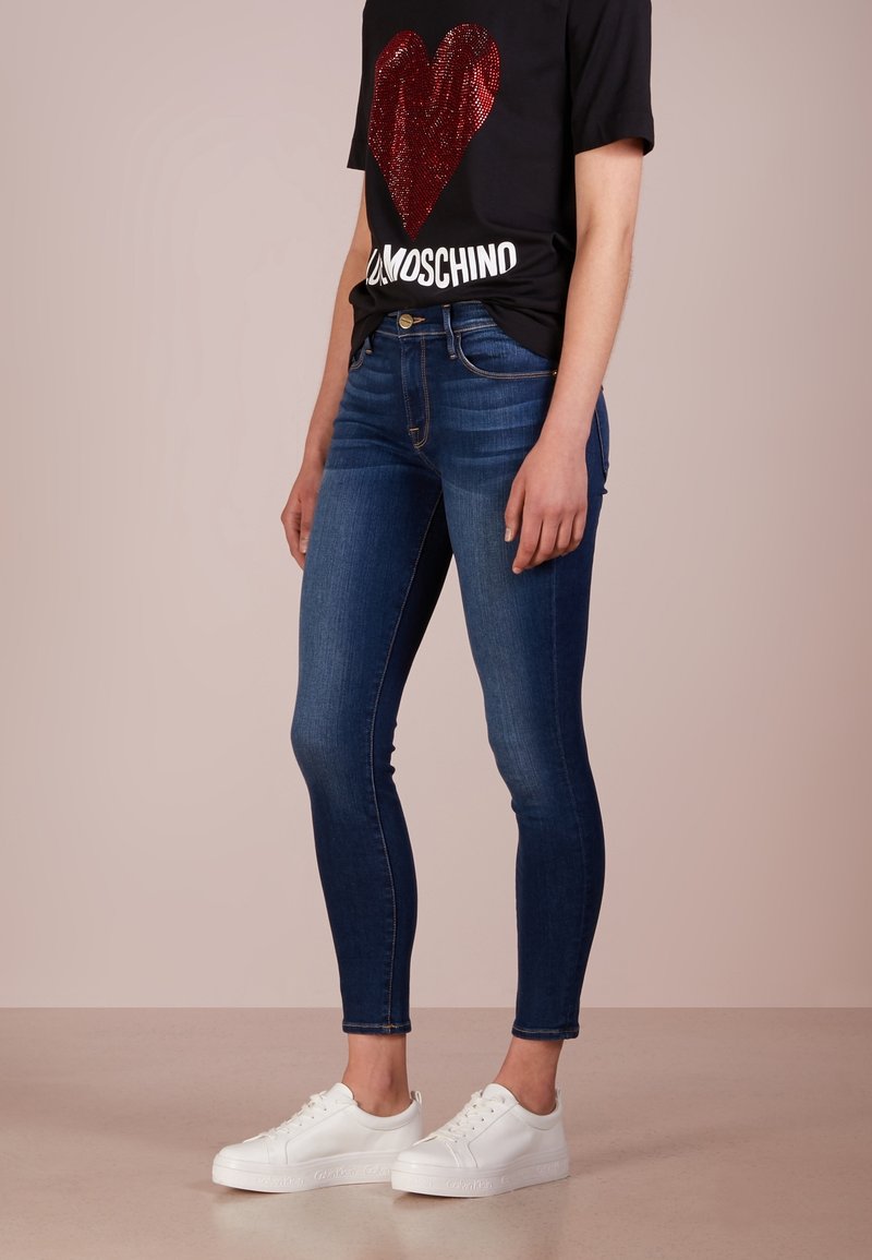 Jeans ajustados en mezclilla azul oscuro con diseño de cinco bolsillos, combinados con una camiseta negra con un corazón de lentejuelas rojas y letras blancas. Zapatillas blancas.