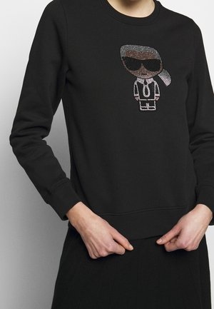 Sweatshirt noir avec un personnage de dessin animé ludique orné de strass, portant des lunettes de soleil et un costume sur le devant.