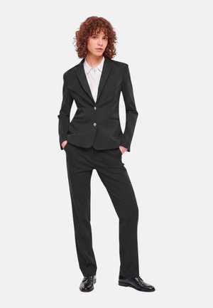 Persona con capelli ricci che indossa un abito nero aderente con camicia bianca con colletto e scarpe nere lucide, in piedi con le mani in tasca.