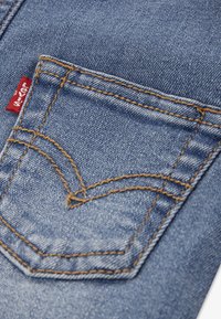 Υφασμα ανοιχτού μπλε τζιν με μικρή πίσω τσέπη και ραφές αντίθετου χρώματος. Εμφανές κόκκινο ετικετάκι μάρκας κοντά στο πάνω μέρος της τσέπης.