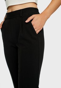 Pantalon noir ajusté avec une texture lisse, une coupe slim, des poches avant et une taille mi-haute.