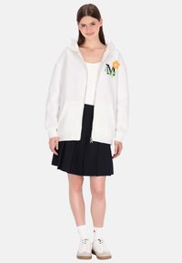 Fehér cipzáras kapucnis pulóver virágos hímzéssel, polár anyagból, állítható húzózsinóros kapucnival, első zsebekkel, tengerészkék pliszírozott szoknyával párosítva.