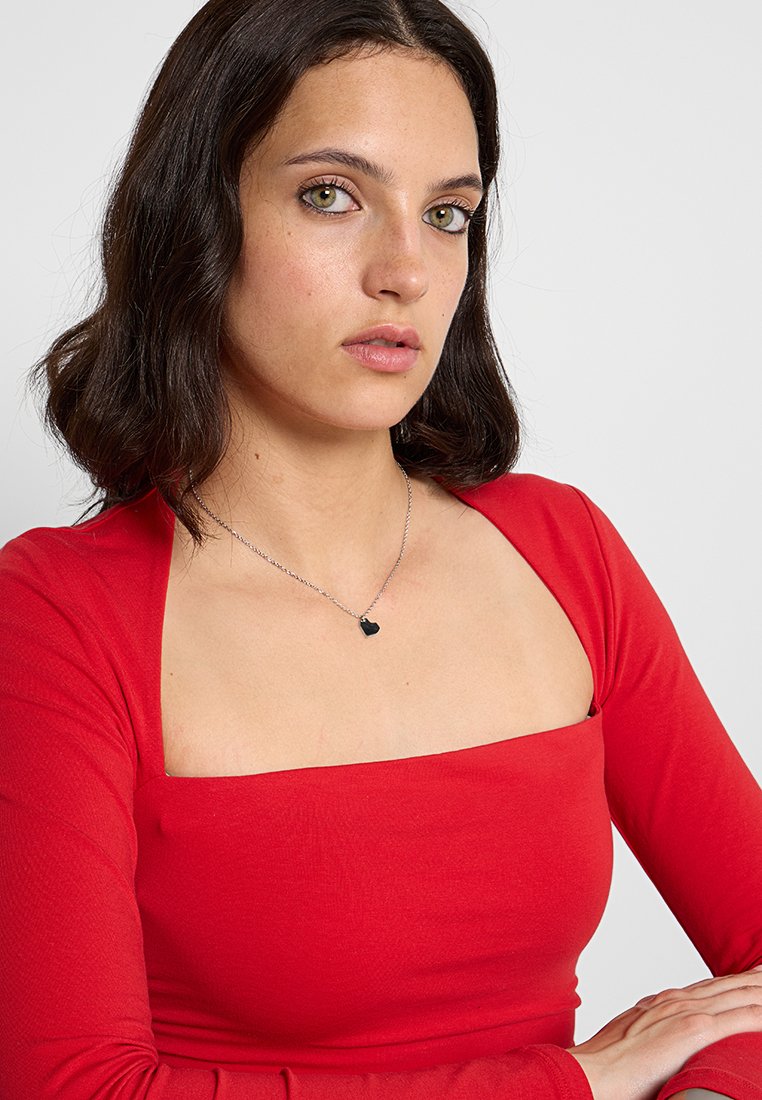 Rotes langärmliges Oberteil mit quadratischem Ausschnitt, kombiniert mit einer silbernen Kettenhalskette, die einen kleinen schwarzen Herzanhänger trägt.