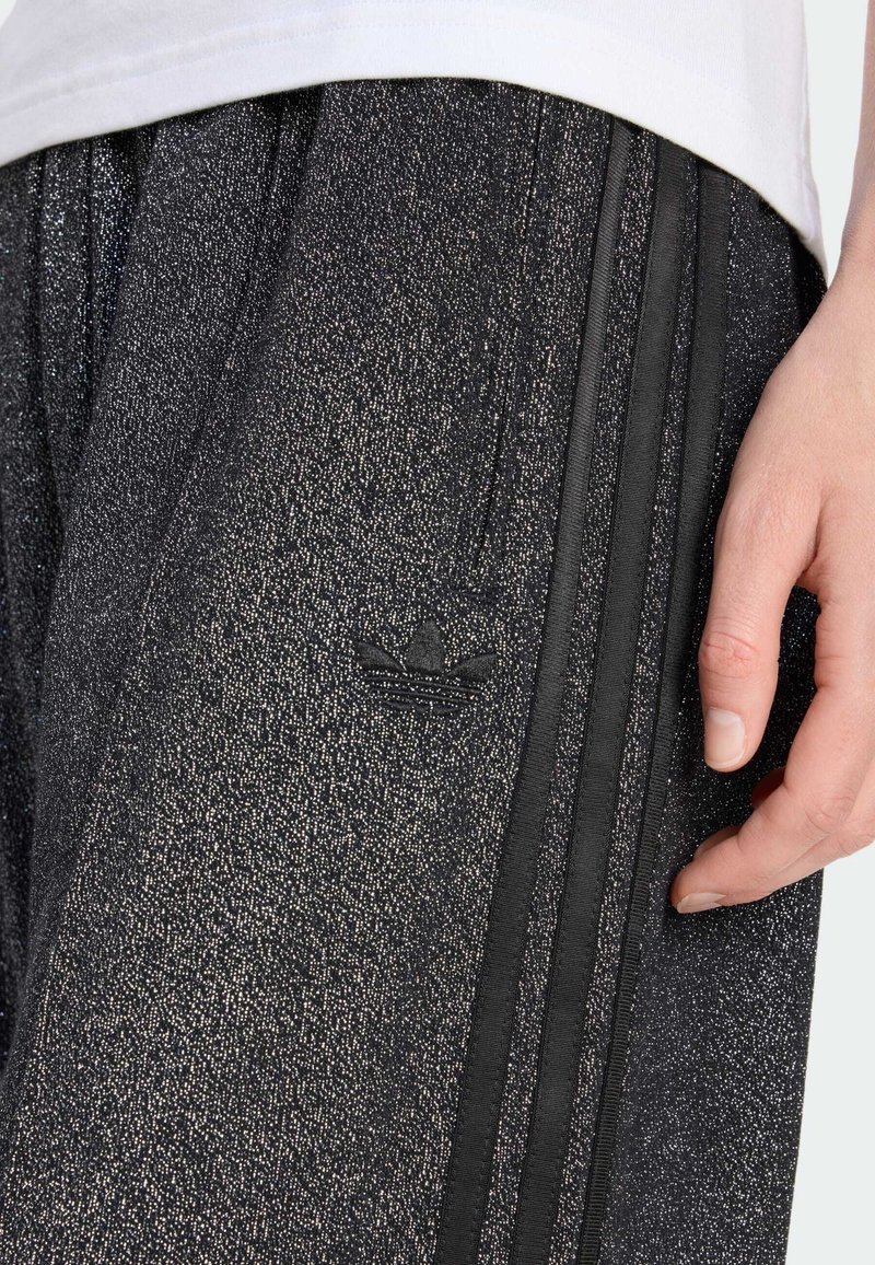 Un pantalon de sport noir avec une finition texturée et scintillante présente trois rayures verticales noires et un logo en relief sur le côté gauche.