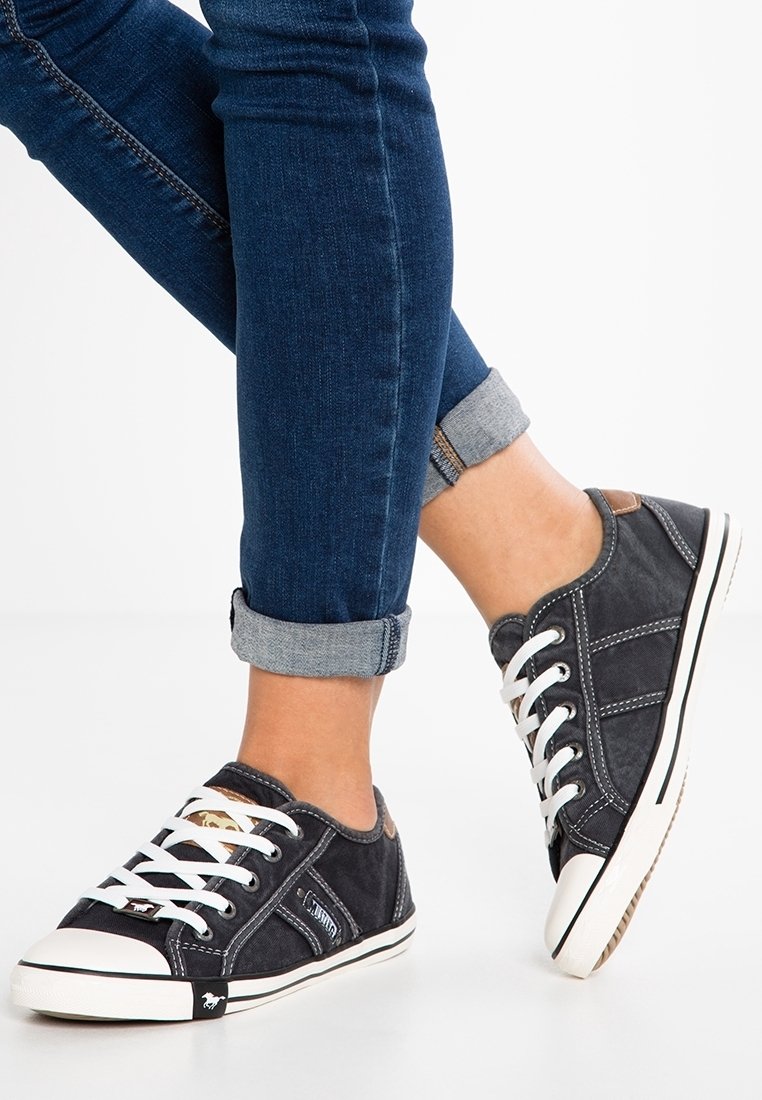 Osoba oblečená v tmavě modrých vyhrnutých džínách a černých plátěných teniskách s bílými tkaničkami a gumovými podrážkami, stojící na bílé podlaze.